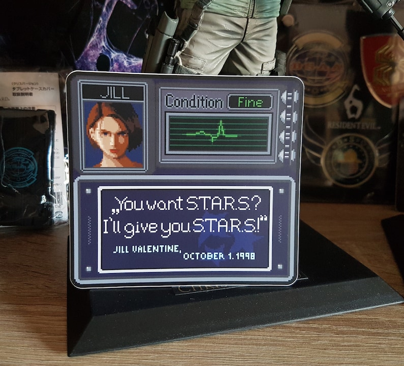 May include: A blue and white sticker with a pixelated image of a woman with the text "JILL" above her. The sticker also has a pixelated image of a computer screen with the text "Condition Fine" and a green line graph. The bottom of the sticker has a pixelated image of a computer screen with the text "You want S.T.A.R.S? I'll give you STARS!" and the text "JILL VALENTINE, OCTOBER 1, 1998".