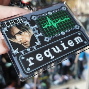 May include: A rectangular keychain with a pixelated design. The top section features the name "LEON" above a portrait. The right side shows a green heartbeat monitor on a grid. The bottom reads "requiem" in white letters.