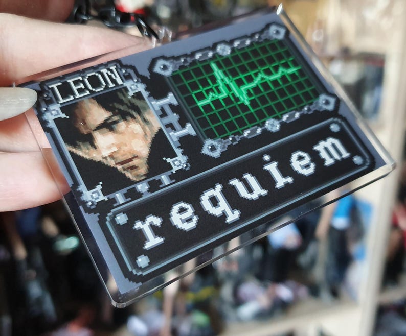 May include: A rectangular keychain with a pixelated design. The top section features the name "LEON" above a portrait. The right side shows a green heartbeat monitor on a grid. The bottom reads "requiem" in white letters.