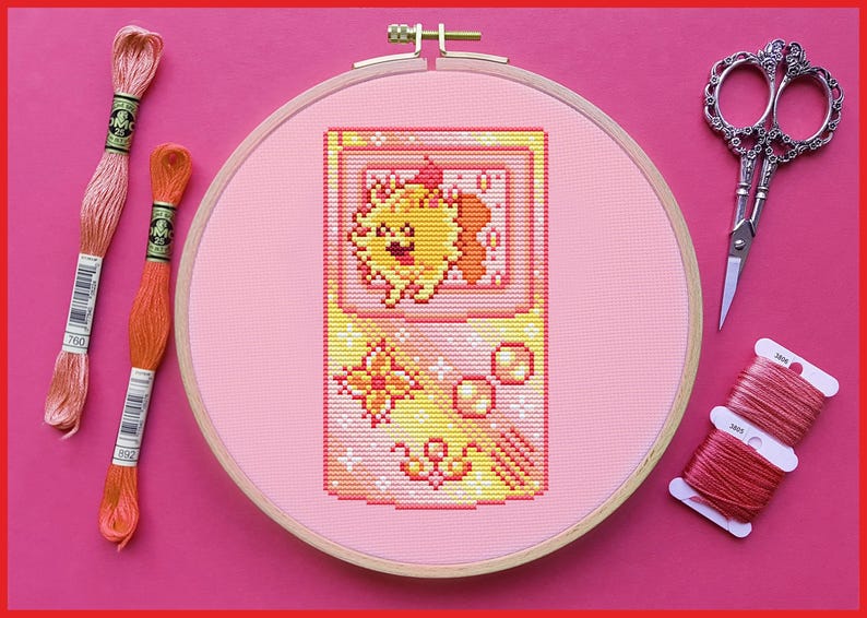 May include: A cross-stitch project featuring a retro video game design with a smiling character. The embroidery is set in a wooden hoop, surrounded by embroidery floss and decorative scissors. The color palette includes pink, yellow, and orange.