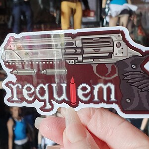 以下が含まれることがあります： 白い文字で「requiem」という言葉が入った、ピクセル化されたリボルバーのデザインのステッカー。銃は銀色とグレーで、濃い赤色の背景に、赤い弾丸が付いています。ステッカーには白い縁取りがあります。