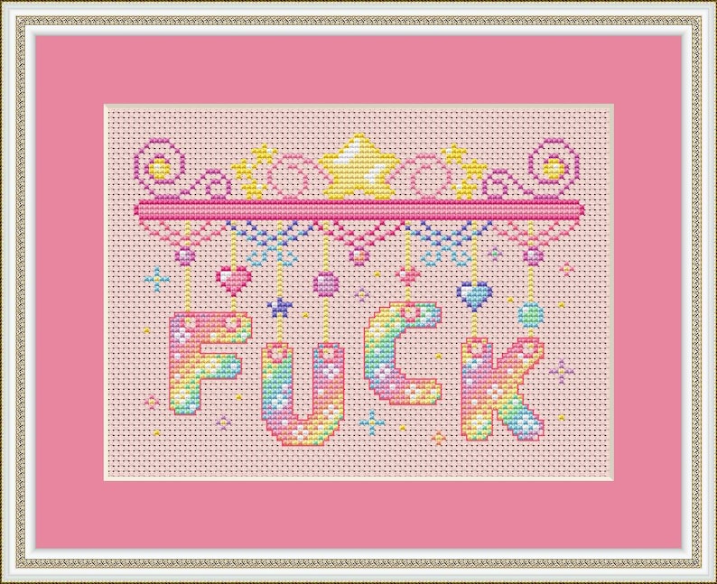 May include: Cross-stitch artwork in a pink frame. The design spells out "FUCK" in rainbow hues, with decorative elements above and below. The background is a light pink fabric, typical of cross-stitch.