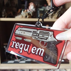 May include: A rectangular keychain with a pixelated gun design on a red background. The word "requiem" is written in white pixelated letters. The keychain is attached to a black metal chain.