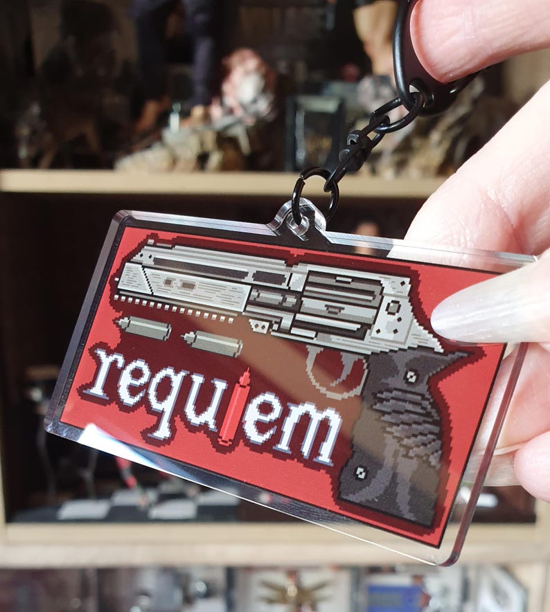 May include: A rectangular keychain with a pixelated gun design on a red background. The word "requiem" is written in white pixelated letters. The keychain is attached to a black metal chain.