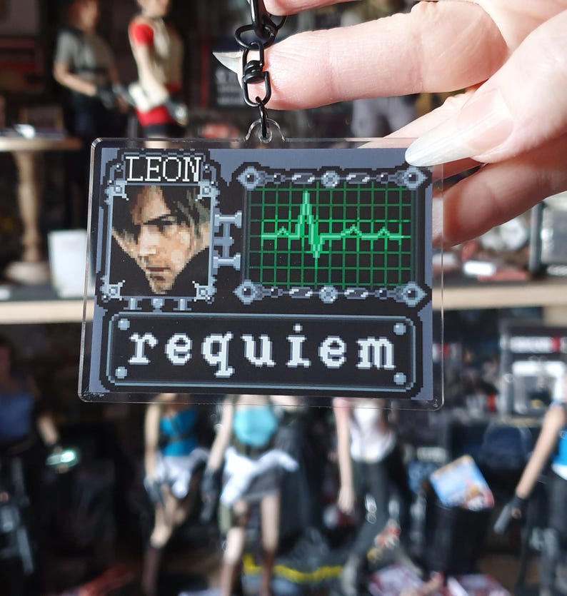 May include: A clear acrylic keychain featuring a pixelated design. The keychain has a portrait of a man, the name "LEON", a green heartbeat monitor, and the word "requiem". The keychain is held by a black metal clasp.