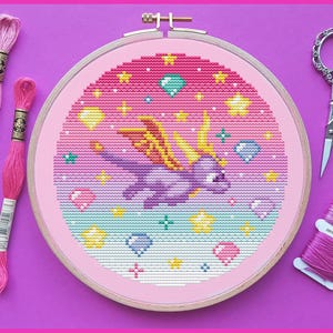 Puede incluir: Un proyecto de punto de cruz terminado con un dragón morado con alas amarillas, rodeado de estrellas y gemas, en un bastidor rosa. Hilos de bordar rosas y tijeras plateadas sobre un fondo morado.