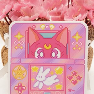 May include: A pink and purple pixel art sticker featuring a cat with a crescent moon on its forehead, a white rabbit, and stars. The sticker is shaped like a handheld video game console.