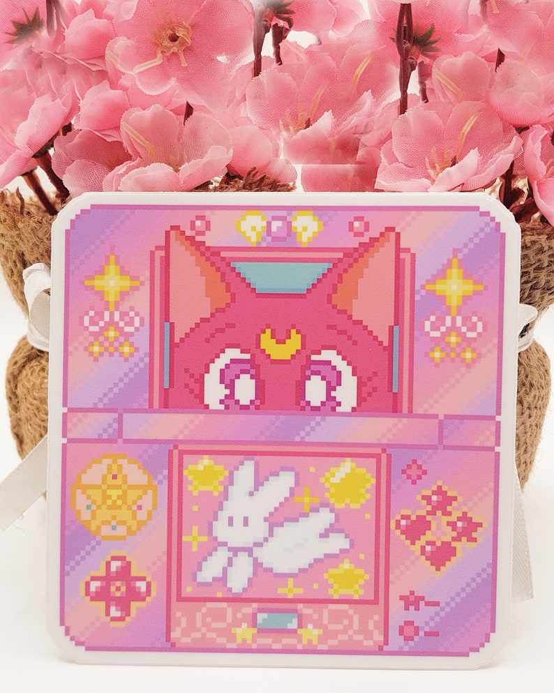 May include: A pink and purple pixel art sticker featuring a cat with a crescent moon on its forehead, a white rabbit, and stars. The sticker is shaped like a handheld video game console.