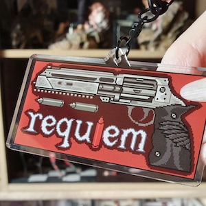 May include: A rectangular keychain with a pixelated gun design on a red background. The word "requiem" is written in white pixelated letters. The keychain is attached to a black metal clasp.