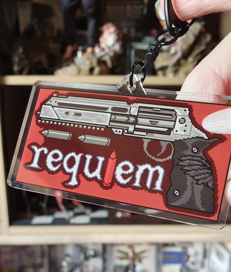May include: A rectangular keychain with a pixelated gun design on a red background. The word "requiem" is written in white pixelated letters. The keychain is attached to a black metal clasp.