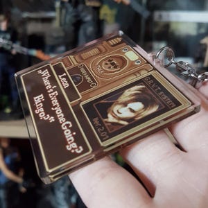 May include: A clear acrylic keychain featuring a brown and tan design with the text "Where's Everyone Going? Bingo?" and "Leon". The keychain also includes a portrait and the text "G.R.V.T. SYSTEM".