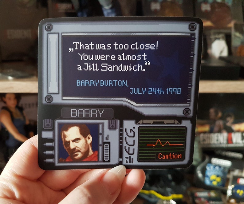 K&ouml;nnte beinhalten: Ein schwarz-wei&szlig;er Aufkleber mit einem pixeligen Bild eines Videospielcharakters. Der Text auf dem Aufkleber lautet: &bdquo;Das war knapp! Du warst fast ein Jill-Sandwich.&ldquo; Barry Burton, 24. Juli 1998. Der Aufkleber enth&auml;lt auch ein pixeliges Bild eines Herzfrequenzmonitors mit dem Wort &bdquo;Vorsicht&ldquo; darunter.