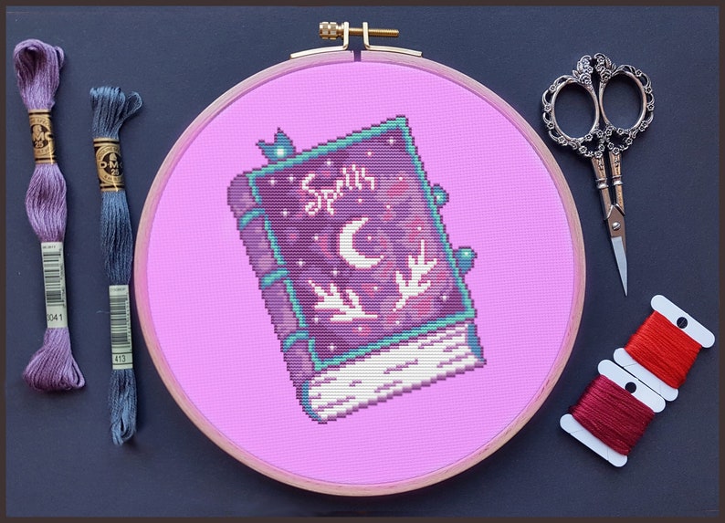 Puede incluir: Un proyecto de punto de cruz terminado en un aro de bordado rosa. El dise&ntilde;o es un libro morado con una cubierta de cielo nocturno estrellado y la palabra "Spells" escrita en la parte delantera. El libro est&aacute; rodeado por un borde turquesa.
