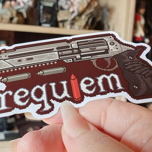 以下が含まれることがあります： 茶色とグレーの色合いでピクセル化された拳銃のデザインのステッカー。白いブロックレターで「requiem」という文字が書かれています。ステッカーには赤い弾丸が含まれています。背景はぼやけています。