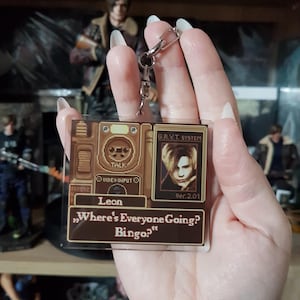 May include: A clear acrylic keychain featuring a brown design with the text "Leon" and the quote "Where's Everyone Going? Bingo." The keychain includes a portrait and a silver chain.