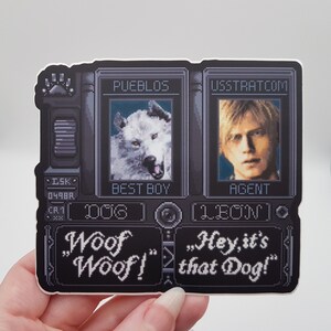 May include: A black and white pixelated sticker with two character portraits. The left portrait is of a white wolf with the text "PUEBLOS" and "BEST BOY". The right portrait is of a man with the text "USSTRATCOM" and "AGENT". The text "Woof Woof!" and "Hey, it's that Dog!" is below the portraits.