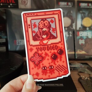 以下が含まれることがあります： 画面にゾンビのキャラクターが表示された、赤と白のピクセル化されたゲームボーイカラーのステッカー。画面には「YOU DIED」と表示されています。