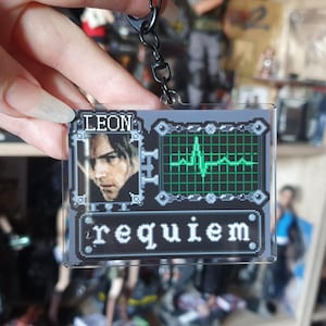 May include: A clear acrylic keychain featuring a pixelated design with the name "LEON" and the word "requiem". The keychain includes a portrait and a green heartbeat monitor graphic. Attached to a black chain.