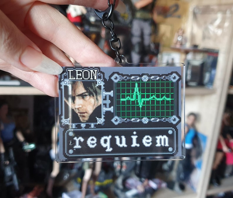 May include: A clear acrylic keychain featuring a pixelated design with the name "LEON" and the word "requiem". The keychain includes a portrait and a green heartbeat monitor graphic. Attached to a black chain.