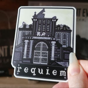 May include: A grayscale sticker featuring a pixelated illustration of a building with the text "Raccoon Police" above a large gate. Below the building is the word "requiem" in white letters on a black background.