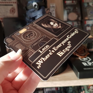 Peut inclure: Un autocollant de jeu vid&eacute;o de couleur marron et noir avec un personnage du jeu Resident Evil 4. L'autocollant repr&eacute;sente un personnage nomm&eacute; Leon et le texte "Where's Everyone Going? Bingo?"