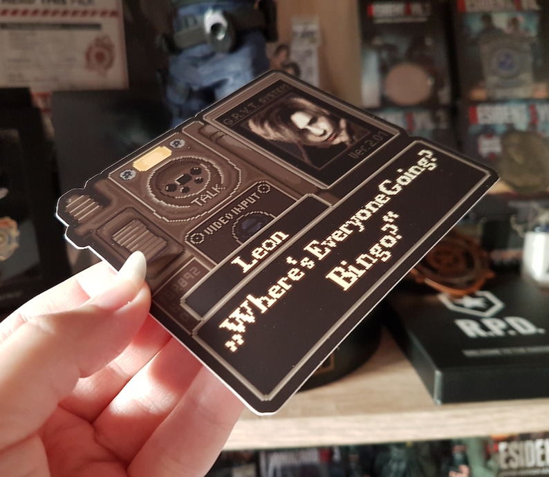 Peut inclure: Un autocollant de jeu vid&eacute;o de couleur marron et noir avec un personnage du jeu Resident Evil 4. L'autocollant repr&eacute;sente un personnage nomm&eacute; Leon et le texte "Where's Everyone Going? Bingo?"