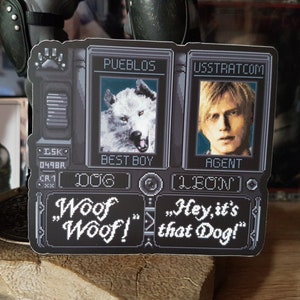 May include: A black and white sticker with a pixelated image of a wolf and a man. The sticker has the text "PUEBLOS" and "USSTRATCOM" above the images. The text "BEST BOY" and "AGENT" are below the images. The text "[DOG]" and "[LEON]" are below the text "BEST BOY" and "AGENT". The text "Woof "Woof!" and "Hey, it's that Dog!" are at the bottom of the sticker.