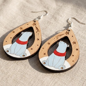 May include: A pair of teardrop-shaped wooden earrings featuring a light blue polar bear wearing a red scarf. The bear sits on a white snowy patch within the cutout, surrounded by engraved stars and a shooting star on the natural wood frame.