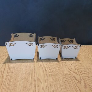 May include: Three wooden boxes with a white painted interior and brown exterior. The boxes have a decorative design of black swirls on the top and bottom edges.