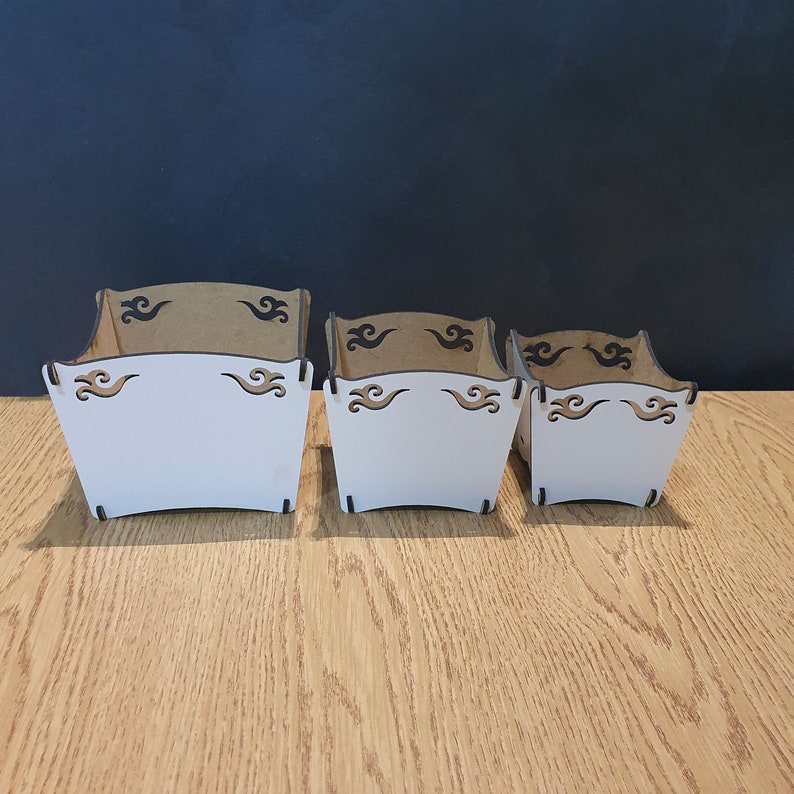 May include: Three wooden boxes with a white painted interior and brown exterior. The boxes have a decorative design of black swirls on the top and bottom edges.