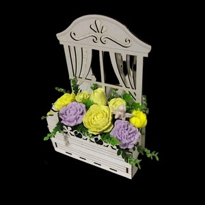 May include: A white wooden window box with a window frame and a floral arrangement of yellow and purple soap flowers. The flowers are arranged in a box with green leaves.