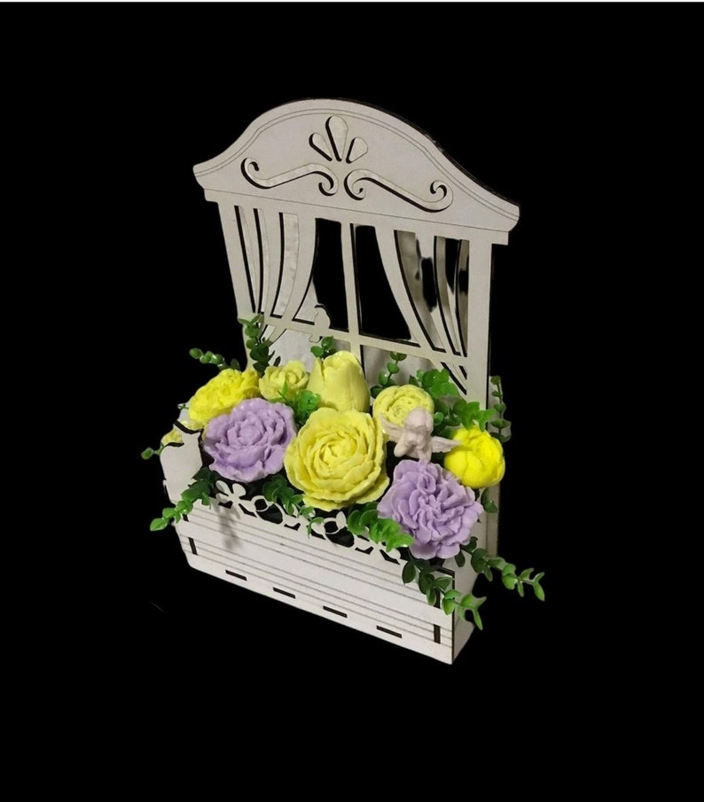 May include: A white wooden window box with a window frame and a floral arrangement of yellow and purple soap flowers. The flowers are arranged in a box with green leaves.