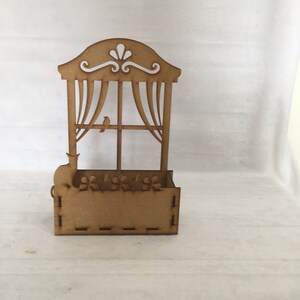 May include: A wooden cutout of a window with curtains, a bird perched on the windowsill, and a cat sitting on a planter box below. The planter box has a decorative design of flowers.