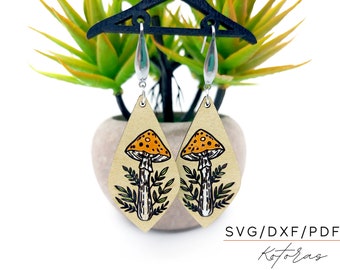 May include: A pair of teardrop-shaped earrings with a mushroom design. The earrings feature a beige background with a black and white mushroom illustration and orange spots on the cap. The earrings are hanging from silver hooks. Text on the image reads "SVG/DXF/PDF" and "Kotoras".