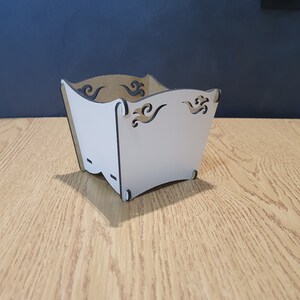 May include: White cardboard box with decorative cutouts in the shape of waves. The box is empty and sitting on a wooden surface.