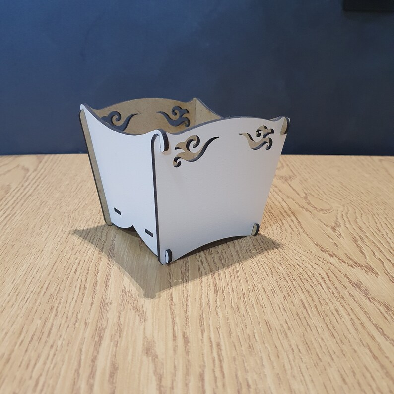 May include: White cardboard box with decorative cutouts in the shape of waves. The box is empty and sitting on a wooden surface.