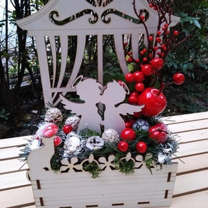 May include: A white wooden Christmas decoration with a window-like design, featuring two angels holding a key, surrounded by red berries, pine cones, and greenery.