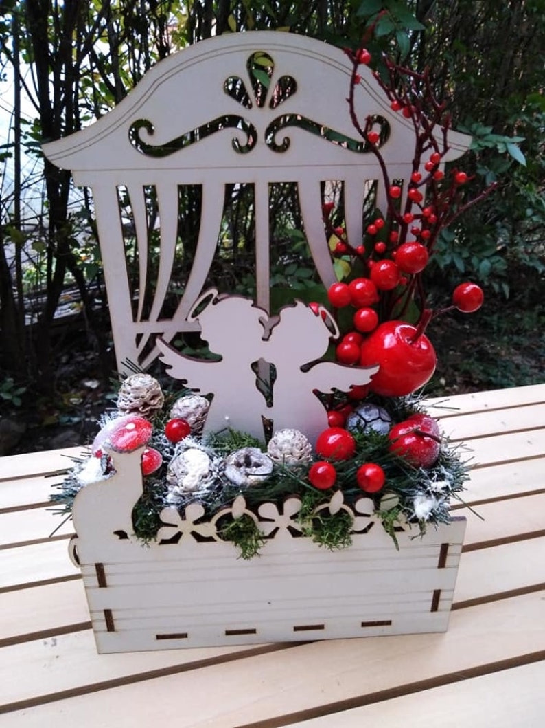 May include: A white wooden Christmas decoration with a window-like design, featuring two angels holding a key, surrounded by red berries, pine cones, and greenery.