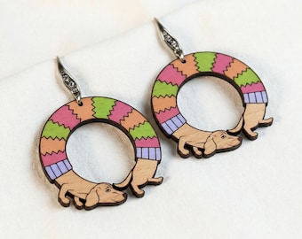 May include: A pair of wooden dachshund earrings. Each earring features a dachshund dog design with a colorful, striped circular sweater in shades of pink, green, orange, and purple. The earrings have silver-colored ear wires.