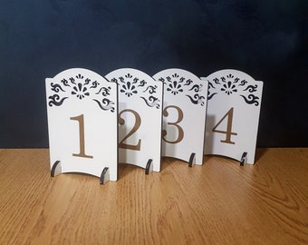 May include: Four white table number signs with a decorative design and gold numbers 1, 2, 3, and 4. The signs are on a wooden surface.