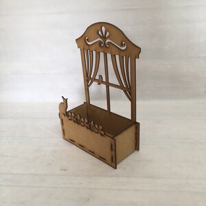 May include: A wooden window planter box with a cutout design of a window with curtains and a bird. A cat silhouette is on the planter ledge. The planter is brown and has a rectangular shape.