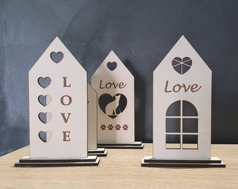 Puede incluir: Tres adornos de madera con forma de casa con la palabra "Love" en cada uno. Un adorno tiene una silueta de gato, otro un corazón y el tercero una ventana.