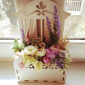 May include: A white wooden planter shaped like a window with a decorative design. The planter is filled with a variety of flowers, including lavender, white roses, and pink flowers.