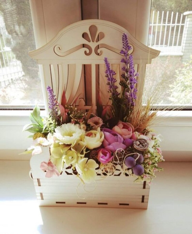 May include: A white wooden planter shaped like a window with a decorative design. The planter is filled with a variety of flowers, including lavender, white roses, and pink flowers.