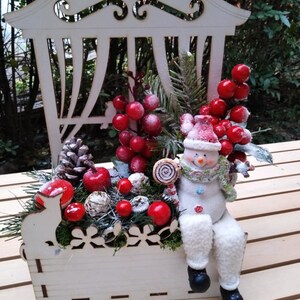 May include: A white wooden window box with a snowman sitting on the edge. The box is decorated with red berries, pine branches, and a small white rabbit figurine. The snowman is holding a candy cane.