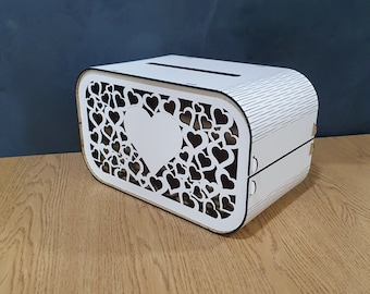 Puede incluir: Caja de madera blanca con un diseño recortado de corazones y un corazón grande en el centro. La caja tiene una ranura en la parte superior para insertar artículos.