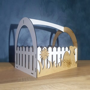 Op de afbeelding: Een houten paasmand in de vorm van een huis met een wit hek, een konijn en een paasei. De mand is versierd met bloemen en gras.