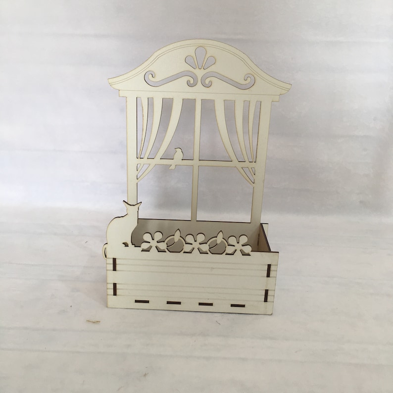 May include: White wooden planter shaped like a window with a cat and bird silhouette. The planter has a decorative top and a window with curtains. The planter has a small box at the bottom with a decorative flower design.