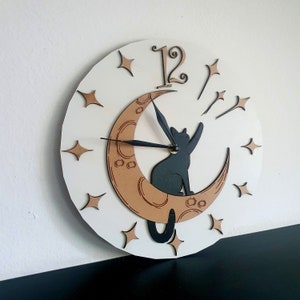 May include: A white clock with black hands and a black cat silhouette sitting on a crescent moon. The moon is brown and has a wood grain texture. The clock face has a white background with black stars and the number 12.