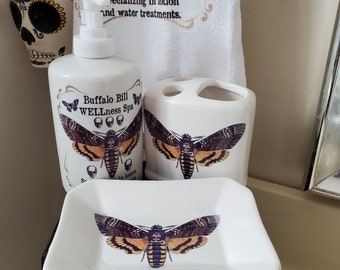 Könnte beinhalten: Ein weißes Badezimmer-Set mit einem braunen und schwarzen Motten-Design. Das Set umfasst eine Seifenschale, einen Zahnbürstenhalter und einen Lotion-Spender. Ein passendes Handtuch mit Motten- und Totenkopf-Grafiken ist ebenfalls sichtbar.